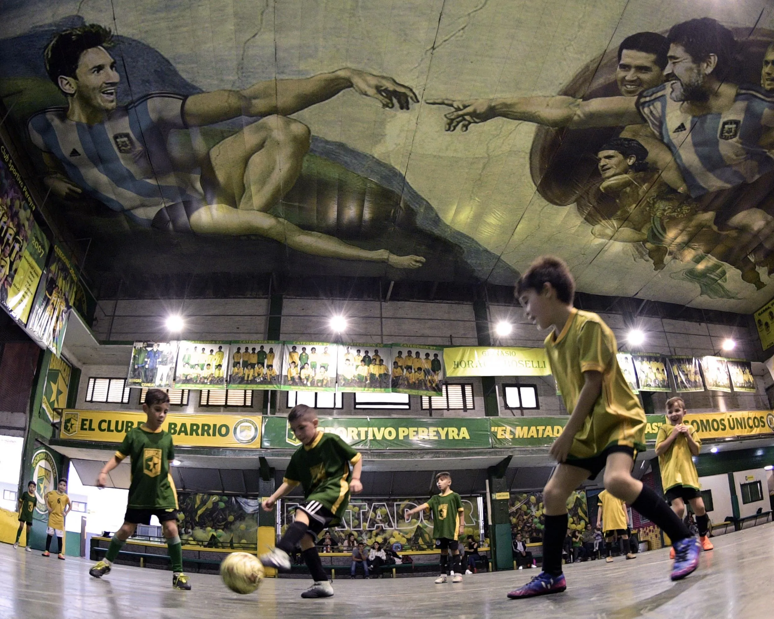 Children play football on an indoor pitch that has a mural on its ceiling of Lionel Messi and Diego Maradona in the style of Michelango’s Creation of Adam.